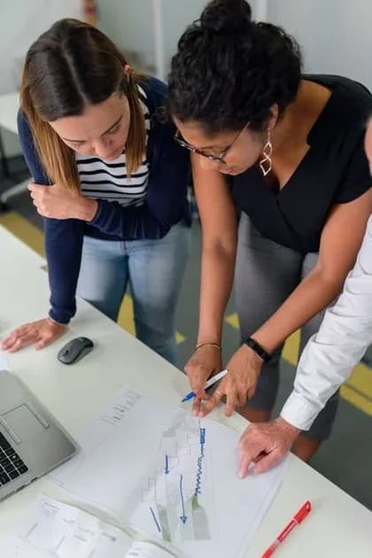 Colleagues gathered around a desk, reviewing documents and pointing at screens during a team planning session.