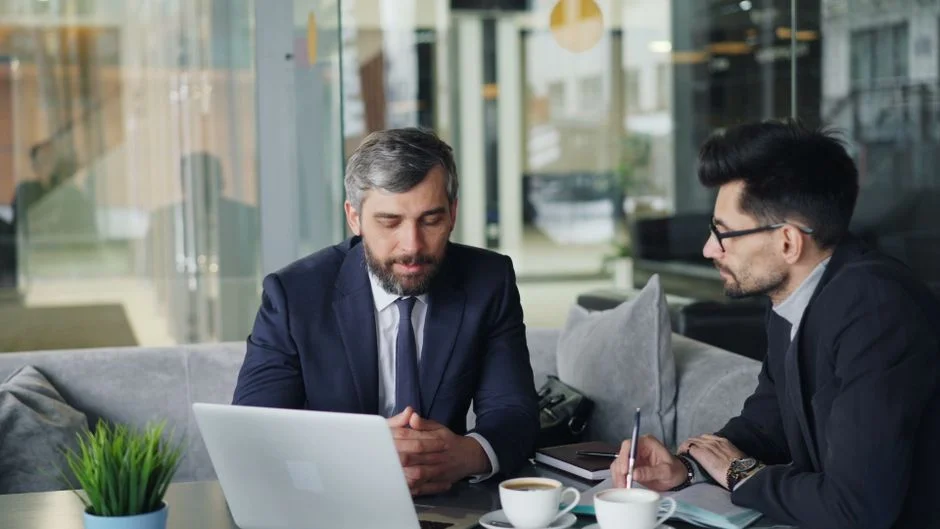 {'answer': 'Two businessmen in suits review work on laptops at a cafe, showcasing revops consulting for B2B startups.'}