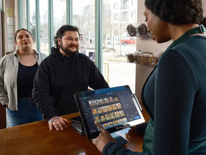 Barista operating a modern touch-screen register while serving two customers at a bright café counter.